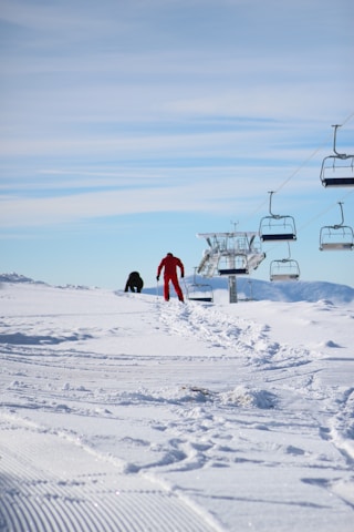 A cheerful family ski sherpa carrying kids' skis and boots from a snowy parking lot toward the ski school lodge.