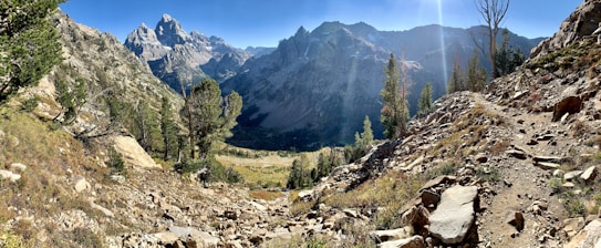 A panoramic view of a rugged mountainous landscape with jagged peaks in the background, surrounded by steep cliffs and rocky terrain. The foreground features a rocky trail winding through sparse vegetation and scattered trees, leading towards a valley. Sunlight filters through, highlighting the textures and casting dynamic shadows.