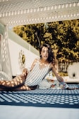 A woman in a striped swimsuit is lounging outdoors under a decorative pergola, which casts intricate shadows over her and the surface she is on. She has wavy hair and wears large hoop earrings. A floral cover-up is draped beside her, and there is a sunlit garden in the background with lush green trees.