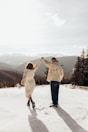 A couple is walking hand in hand on a snowy mountain landscape. The woman is wearing a white dress and holding a small bouquet of flowers, while the man is in a patterned jacket and jeans. They are surrounded by a panoramic view of mountains and trees under a bright, partly cloudy sky.