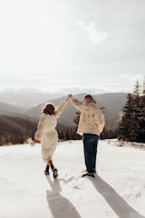A couple is walking hand in hand on a snowy mountain landscape. The woman is wearing a white dress and holding a small bouquet of flowers, while the man is in a patterned jacket and jeans. They are surrounded by a panoramic view of mountains and trees under a bright, partly cloudy sky.