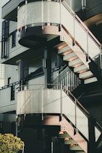 An outdoor spiral staircase with glass railing and metal structure, attached to a modern building with a textured facade. Soft sunlight casts shadows on the stairs and walls.