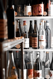 A variety of sake bottles with different labels are arranged on shelves inside a refrigerated display. The bottles vary in color and size, featuring a mix of traditional and modern label designs including characters and symbols.