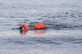 A swimmer is in the water with one arm extended forward, wearing a bright orange flotation device on their back. The water is slightly rippled, indicating motion.
