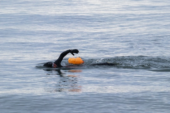 A swimmer wearing a wetsuit is in open water, performing a freestyle stroke. The swimmer accompanies a bright orange buoy floating beside them.