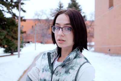 A woman with dark hair and glasses stands outdoors in a snowy setting near a brick building and a path lined with trees. She is wearing a patterned, sleeveless puff jacket over a long-sleeve shirt.