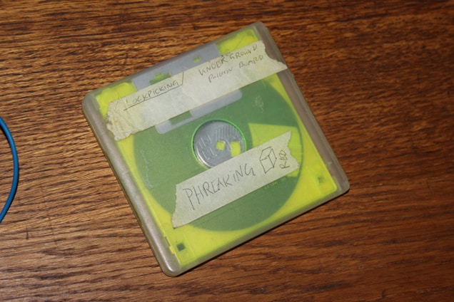 A vintage floppy disk case is placed on a wooden surface. The case is transparent with a yellow liner visible inside. Handwritten labels are attached to the case with words such as 'LOCKPICKING' and 'PHREAKING'.