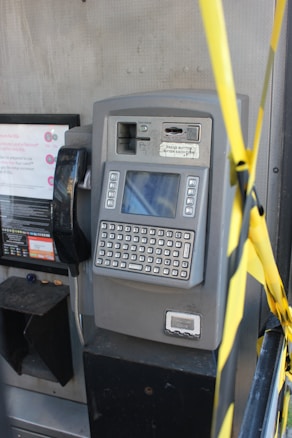 A gray public payphone with a keypad and screen is mounted on a metal wall. There are coin slots and a card slot above the keypad. Yellow caution tape is partially visible on the right side of the image.