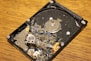 A damaged hard disk drive with broken platters scattered across its surface, showing multiple shards and debris while resting on a wooden surface.