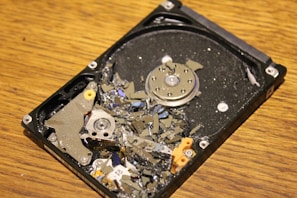 A damaged hard disk drive with broken platters scattered across its surface, showing multiple shards and debris while resting on a wooden surface.