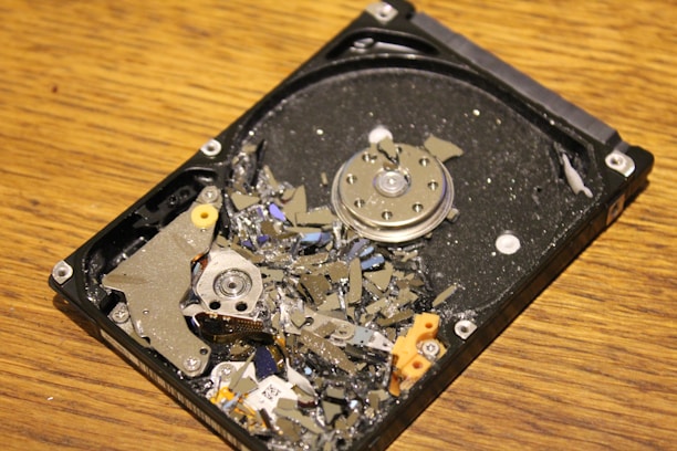 A damaged hard disk drive with broken platters scattered across its surface, showing multiple shards and debris while resting on a wooden surface.