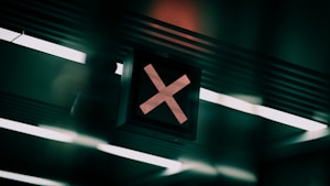 A dark electronic sign with a red illuminated cross symbol, mounted on a ceiling with linear fluorescent lights casting a futuristic and moody glow.