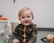 A young child with light hair is smiling while wearing a camouflage-patterned jacket. The child is surrounded by soft and colorful toys in a playroom setting.
