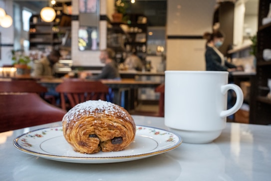 A cozy caf&eacute; scene with a pastry on a decorative plate placed on a table alongside a white ceramic mug. In the background, there are blurred figures of people seated and working, and a barista behind the counter, creating a warm and inviting atmosphere. The caf&eacute; is decorated with shelves featuring various items and planters.