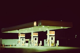 A dimly lit gas station with a canopy displaying the Shell logo, illuminated against a dark night sky. The station features multiple fuel pumps with signage promoting 'Combustibles de Calidad', and the area appears deserted with no vehicles or people present.