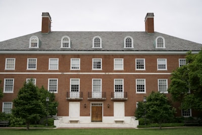 A symmetrical, multi-story brick building with multiple windows and dormer windows on the roof. Two chimneys are visible at either end. The entrance features a wooden door with large steps leading to it, flanked by greenery and lampposts.