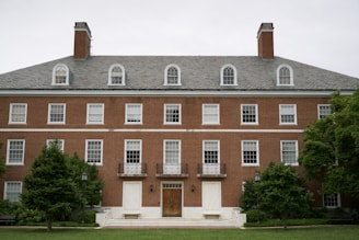A symmetrical, multi-story brick building with multiple windows and dormer windows on the roof. Two chimneys are visible at either end. The entrance features a wooden door with large steps leading to it, flanked by greenery and lampposts.