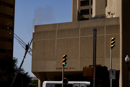 An urban scene featuring traffic lights at an intersection, part of a beige building with a clearance sign, and a pole with wires possibly damaged or leaning. A lamp post and the top of a vehicle are also visible.