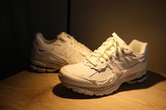 A pair of light-colored athletic shoes with a textured design are placed on a wooden surface. The lighting is warm, casting soft shadows on the surface.