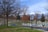 A cemetery with numerous white and gray headstones is situated on neatly maintained green grass. Leafless trees are scattered throughout the area, and a winding paved road divides the cemetery. The sky is overcast with shades of blue and gray, adding to the solemn atmosphere.