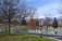A cemetery with numerous white and gray headstones is situated on neatly maintained green grass. Leafless trees are scattered throughout the area, and a winding paved road divides the cemetery. The sky is overcast with shades of blue and gray, adding to the solemn atmosphere.