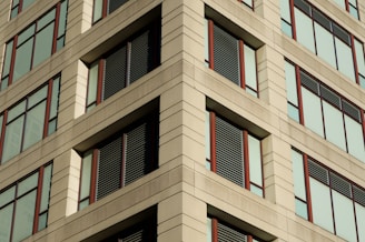 A building facade featuring a symmetrical arrangement of large windows with brown frames. The structure is composed of light-colored stone or concrete panels, creating a pattern with horizontal and vertical lines. Some of the windows have dark slats or panels for shading or as architectural elements.