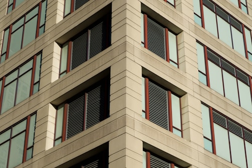 A building facade featuring a symmetrical arrangement of large windows with brown frames. The structure is composed of light-colored stone or concrete panels, creating a pattern with horizontal and vertical lines. Some of the windows have dark slats or panels for shading or as architectural elements.