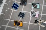 A collection of enamel pins arranged on a gray plaid fabric. The pins are designed to resemble cameras and software icons, including a blue rectangle with 'Ps' for Photoshop and a darker rectangle with 'Lr' for Lightroom. The camera pins come in various colors such as yellow, pink, and metallic hues.