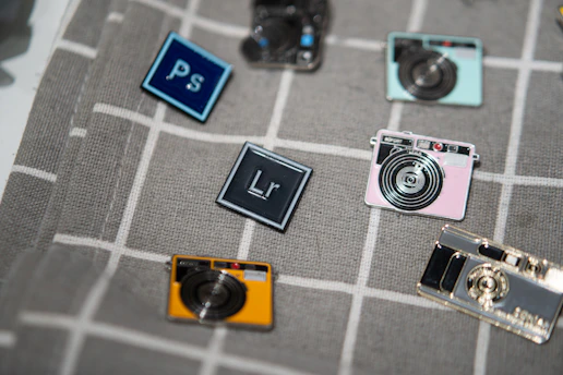 A collection of enamel pins arranged on a gray plaid fabric. The pins are designed to resemble cameras and software icons, including a blue rectangle with 'Ps' for Photoshop and a darker rectangle with 'Lr' for Lightroom. The camera pins come in various colors such as yellow, pink, and metallic hues.