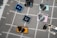 A collection of enamel pins arranged on a gray plaid fabric. The pins are designed to resemble cameras and software icons, including a blue rectangle with 'Ps' for Photoshop and a darker rectangle with 'Lr' for Lightroom. The camera pins come in various colors such as yellow, pink, and metallic hues.