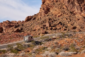 A motorhome travels along a winding road through a desert landscape with rocky hills and sparse vegetation. The reddish-brown rocks dominate the scene, while scattered shrubs grow on the arid ground. The sky is partially cloudy, adding depth to the setting.