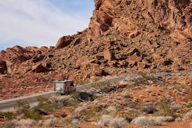 A motorhome travels along a winding road through a desert landscape with rocky hills and sparse vegetation. The reddish-brown rocks dominate the scene, while scattered shrubs grow on the arid ground. The sky is partially cloudy, adding depth to the setting.