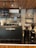 A modern coffee shop interior featuring a black coffee machine with the label 'Cold'N Brew'. To the right are two coffee grinders filled with coffee beans. Shelves in the background hold various glassware and coffee-making equipment. The ceiling is wooden, enhancing the warm ambiance.