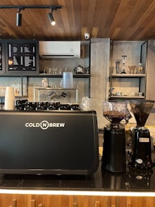 A modern coffee shop interior featuring a black coffee machine with the label 'Cold'N Brew'. To the right are two coffee grinders filled with coffee beans. Shelves in the background hold various glassware and coffee-making equipment. The ceiling is wooden, enhancing the warm ambiance.