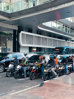 A group of motorcyclists wearing helmets are waiting at an intersection in a bustling city. Several cars are visible behind them, while a modern overpass with glass and metallic elements forms a backdrop. The scene suggests a busy urban environment.