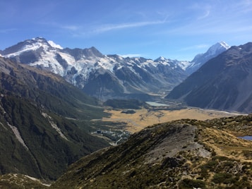 A panoramic view of a mountainous landscape, featuring snow-capped peaks and a deep valley with patches of greenery and a small water body. The sky is clear and blue, adding to the expansive feel of the vista.