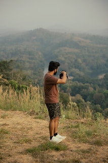 A person stands outdoors in a natural setting, holding a camera up as they take a photo. They are surrounded by tall grass and overlook a vast, hilly landscape covered in dense greenery. The sky appears overcast, creating a subdued atmosphere.