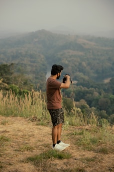 A person stands outdoors in a natural setting, holding a camera up as they take a photo. They are surrounded by tall grass and overlook a vast, hilly landscape covered in dense greenery. The sky appears overcast, creating a subdued atmosphere.