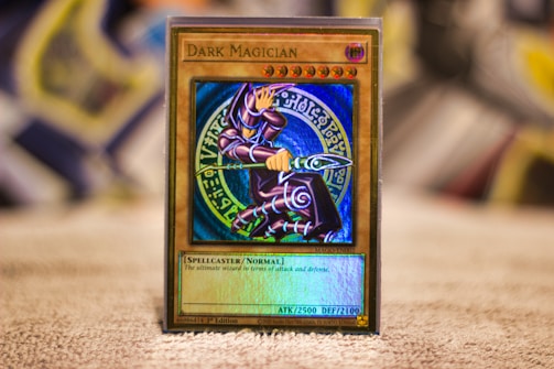 A Yu-Gi-Oh! trading card called 'Dark Magician' is placed upright on a surface. The card features a detailed illustration of a spellcaster in a purple robe, holding a staff and posing dynamically. The background behind the spellcaster is a mystical blue circle with various symbols. The card border is gold, and the text below the image specifies the card's attributes, such as attack and defense points.