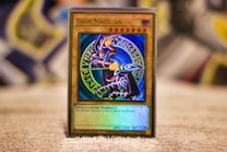 A Yu-Gi-Oh! trading card called 'Dark Magician' is placed upright on a surface. The card features a detailed illustration of a spellcaster in a purple robe, holding a staff and posing dynamically. The background behind the spellcaster is a mystical blue circle with various symbols. The card border is gold, and the text below the image specifies the card's attributes, such as attack and defense points.