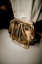 A luxurious gold handbag with a shiny, metallic finish is paired with a chunky gold chain strap. It is set against a textured fabric background, possibly tweed, featuring a checkered pattern and a subtle button accent.