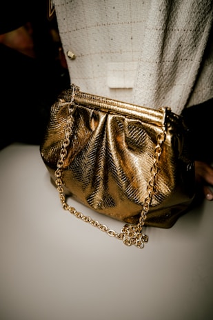 A luxurious gold handbag with a shiny, metallic finish is paired with a chunky gold chain strap. It is set against a textured fabric background, possibly tweed, featuring a checkered pattern and a subtle button accent.