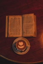 An open book with pages exposed lies on a dark wooden surface beside a cup of coffee. The coffee has a heart-shaped latte art on top. Coffee beans are scattered around the cup's saucer, adding to the cozy ambiance.