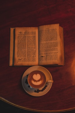 An open book with pages exposed lies on a dark wooden surface beside a cup of coffee. The coffee has a heart-shaped latte art on top. Coffee beans are scattered around the cup's saucer, adding to the cozy ambiance.