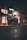 A shopping cart is positioned in the middle of a dimly lit street or sidewalk, partially filled with various items. The background features a softly illuminated building with large windows and some blurred streetlights, giving the scene an urban night-time setting. The atmosphere suggests a feeling of quiet or isolation.