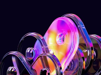 A collection of abstract, translucent shapes with reflective surfaces and bright, vibrant colors. The shapes are rounded and stacked in an overlapping manner against a dark background, creating a futuristic and dynamic appearance.
