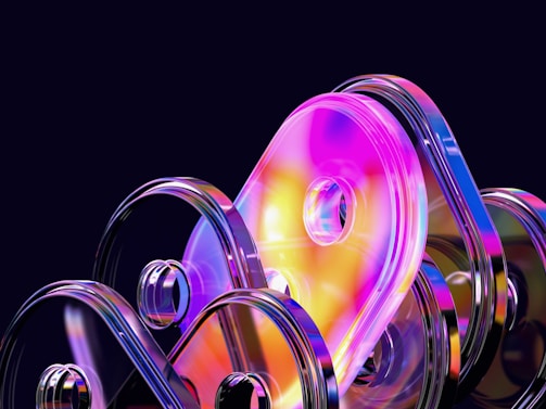A collection of abstract, translucent shapes with reflective surfaces and bright, vibrant colors. The shapes are rounded and stacked in an overlapping manner against a dark background, creating a futuristic and dynamic appearance.