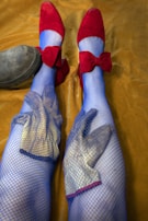 Two legs are adorned in bright blue fishnet stockings, with red shoes featuring large bows. Gloves are inside the stockings, creating a textured appearance. The background is a mustard yellow fabric, and a black boot rests partially visible on the left side.