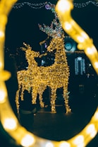 Two reindeer sculptures made of golden lights stand prominently in a festive outdoor setting. Surrounding them are additional decorative lights, and in the background, a clock tower is visible against the dark night sky. The bottom frame is adorned with blurred lights, adding a warm and magical glow to the scene.