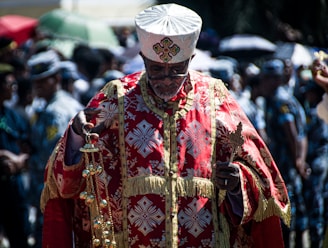 A man dressed in ornate, ceremonial garments with vibrant red and gold patterns stands amidst a crowd. He holds a decorated religious implement in one hand and a censer in the other. The background is blurred, with people using umbrellas for shade.